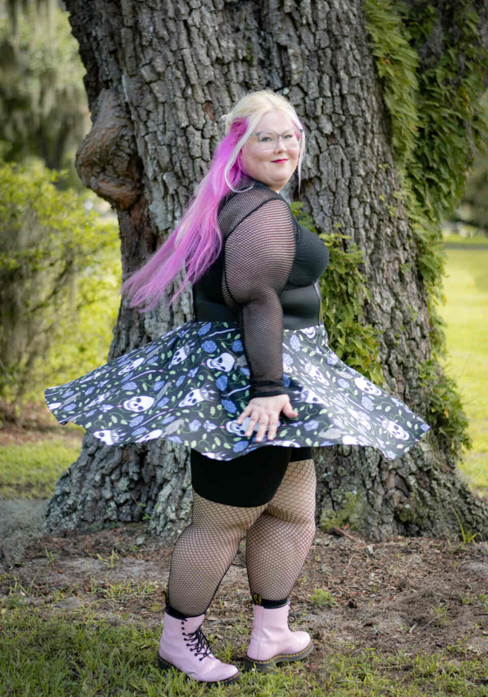 A model wearing a Halloween-themed skirt in an outdoor setting. The model wears a black top, pink boots, and patterned skirt featuring skulls and blue roses on a black background.