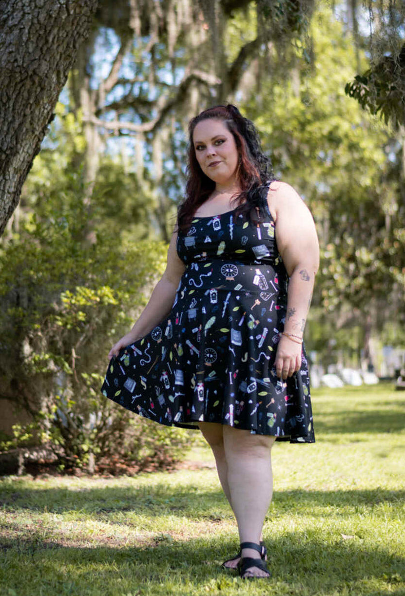 A woman wearing a black sleeveless midi dress with a white and light blue occult-themed graphic print stands in a grassy park area with large trees and hanging moss in the background.