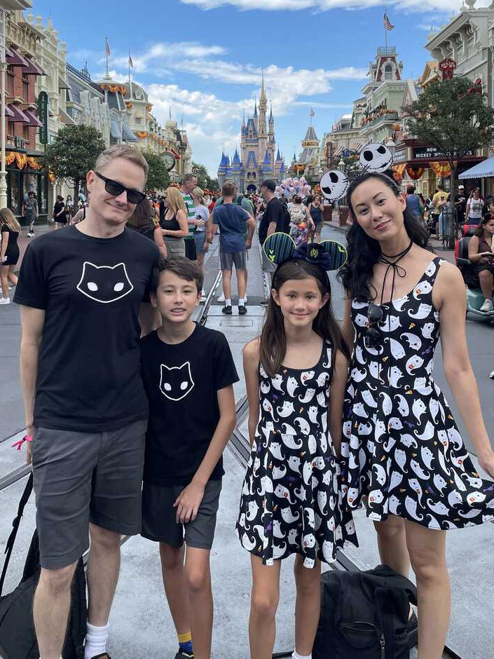A family of four posing on a busy theme park street with a castle in the background. The father and son wear matching black t-shirts with a white cat face graphic, while the mother wears a black and white patterned dress.