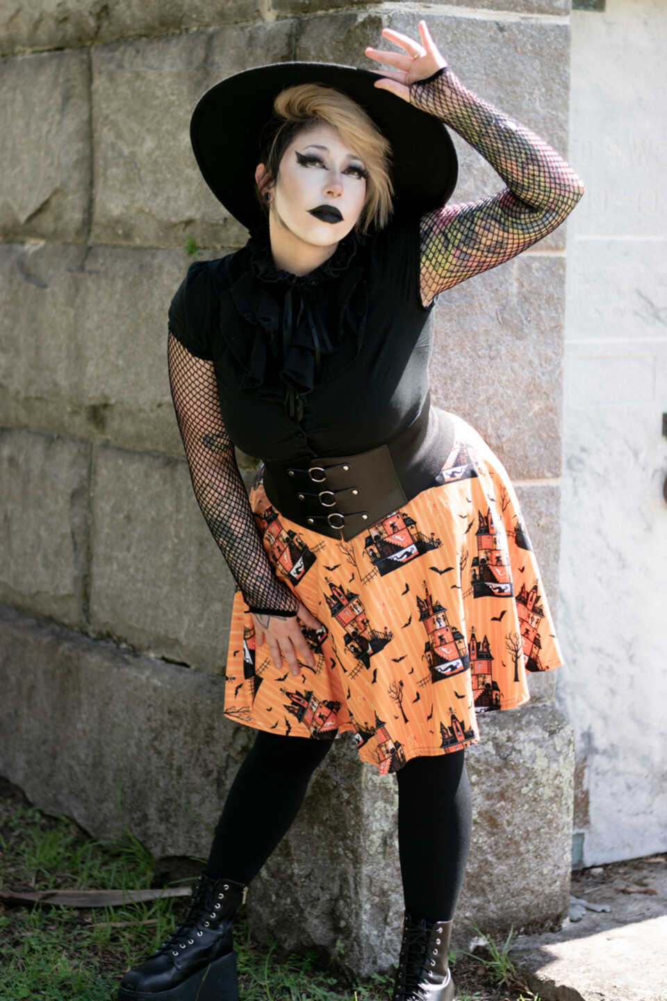 A model wearing a Halloween-themed skirt in an outdoor setting. The model wears a black top, black hat and patterned skirt featuring a haunted house pattern swirling with bats on an orange background.