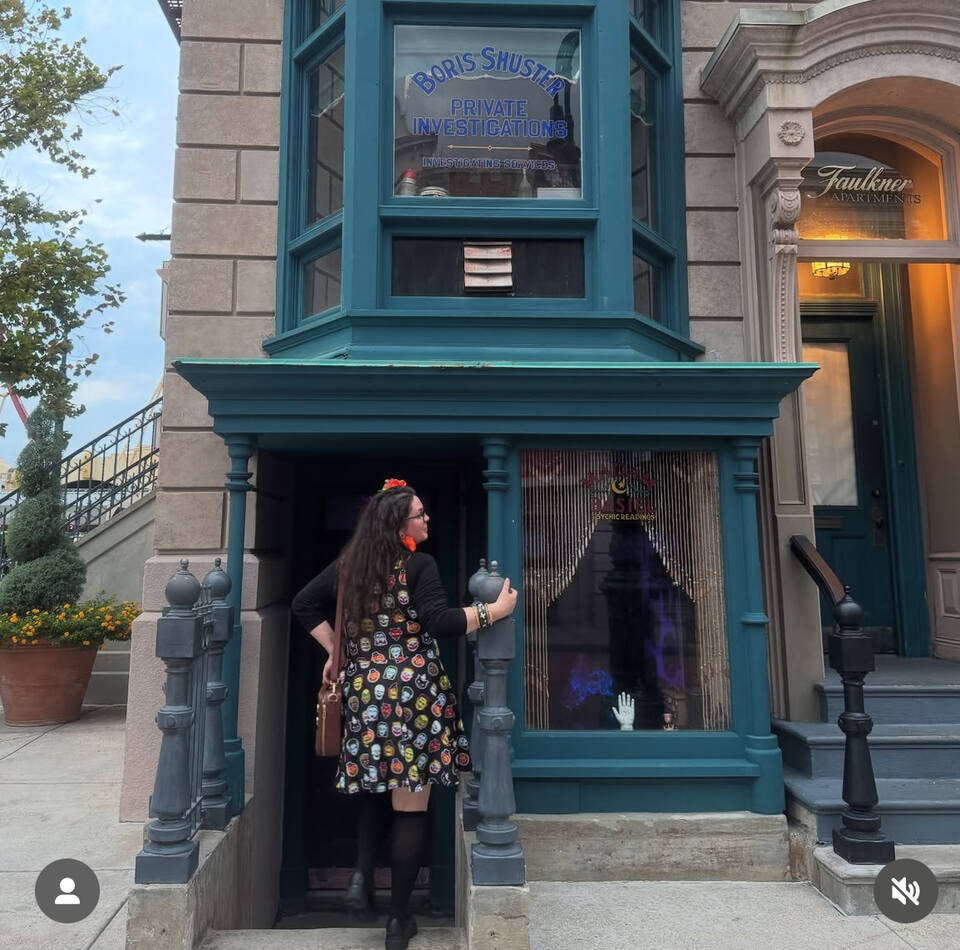 A woman in vintage mask halloween dress stands at the doorway at Universal Studios Florida with teal bayonet windows. The storefront has a sign reading 'Boris Shuster' 
