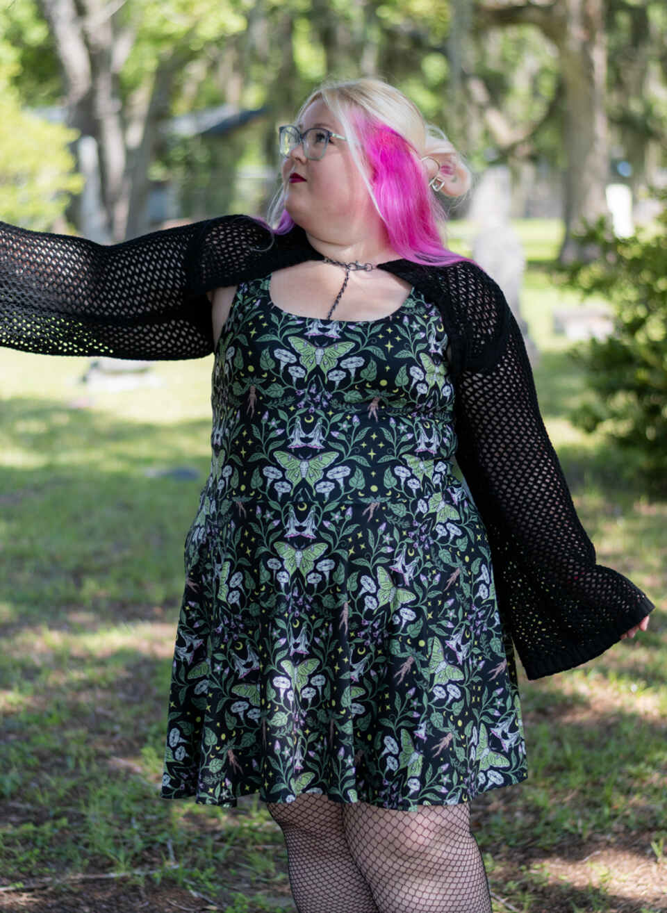 A woman outdoors in a grassy park wearing a dark floral print with luna moth's on a dress with a black open-knit cardigan, fishnet tights, and bright pink hair.