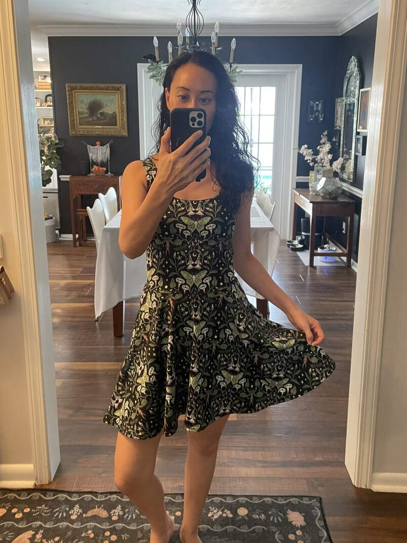 A woman takes a mirror selfie in a home interior, wearing a sleeveless black dress with a light green floral or butterfly-like pattern and a flared skirt.