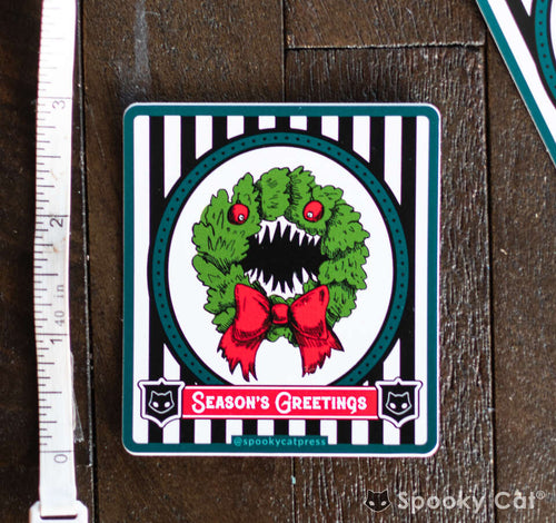 Vinyl sticker of a monster Christmas wreath with sharp teeth and a red bow reading “Season’s Greetings” on a black-and-white striped background.