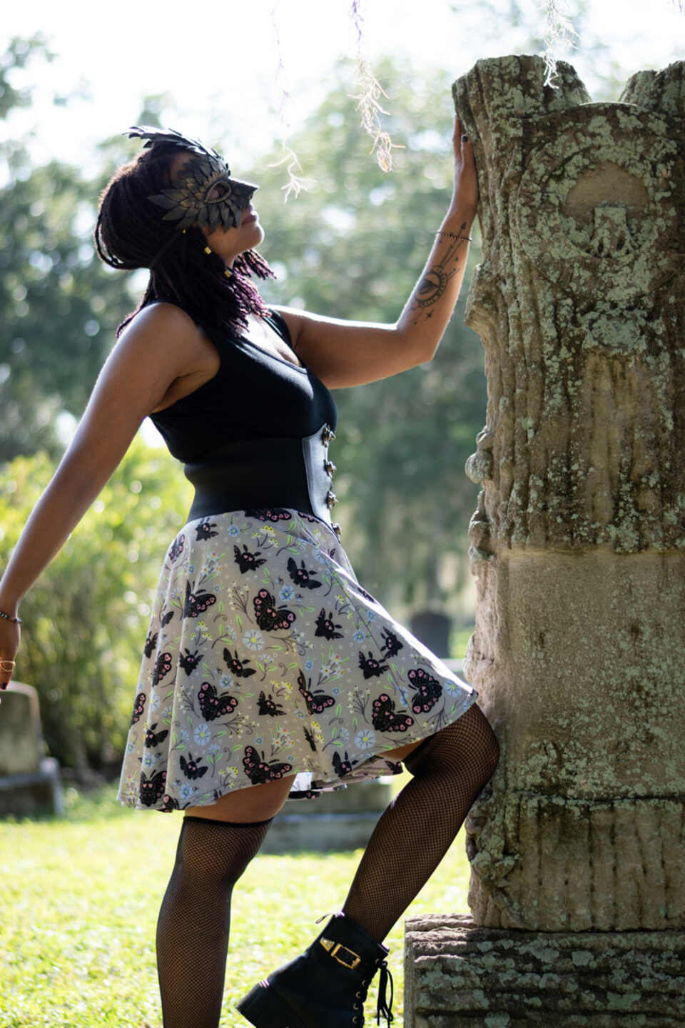 A model wearing a Halloween-themed skirt in an outdoor setting. The model wears a black top, mask, and patterned skirt featuring black mothman with pastel floral flourish on a light grey background.