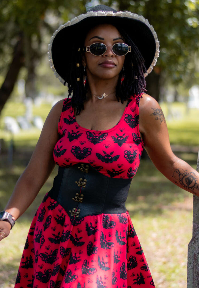 A woman wearing a red and black bat-Mothman Heart Pattern dress with a black corset-style waist, a large black sun hat, and sunglasses, standing outdoors in a park-like setting.