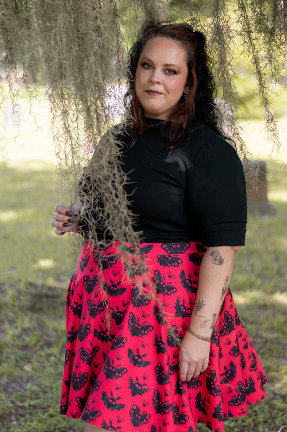 A model wearing a Halloween-themed skirt in an outdoor setting. The model wears a black top, and patterned skirt featuring black heart-shaped mothmen on a red background.