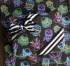 Wrapped gift box with black and white striped ribbon on top of neon-themed gift wrap featuring classic Universal Monsters like Frankenstein, Dracula, Creature from the Black Lagoon, and more. The monsters are illustrated in glowing 80s-style neon colors on a black background