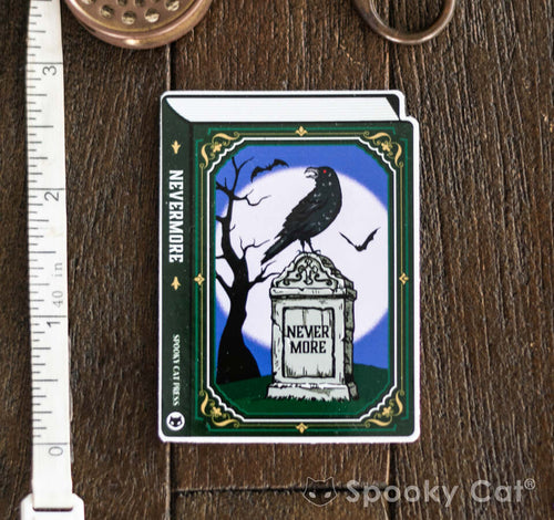 Diecut vinyl sticker shaped like a book with a raven perched on a tombstone that reads “Nevermore” against a moonlit background.