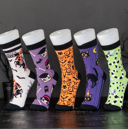 Five pairs of Spooky Cat Press Halloween-themed crew socks displayed on white mannequin legs against a dark gray background. The socks feature spooky designs including a witch, skeletons, bats, a vampire, a cat, and a spider on white, purple, orange, purple, and light green backgrounds.