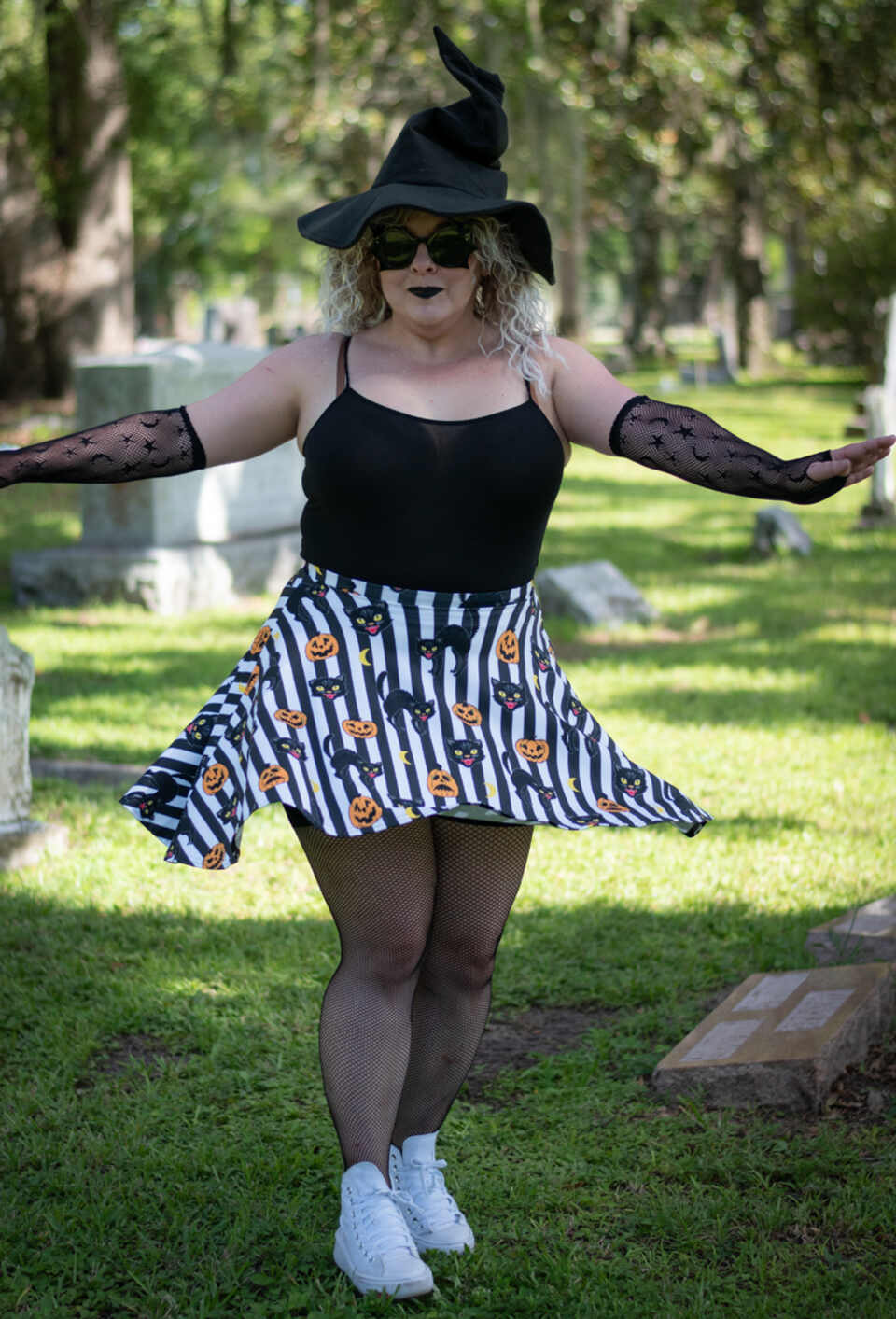 A model wearing a Halloween-themed skirt in an outdoor setting. The model wears a black witch hat and a black-and-white striped skirt featuring orange pumpkins, black cats, and crescent moons.