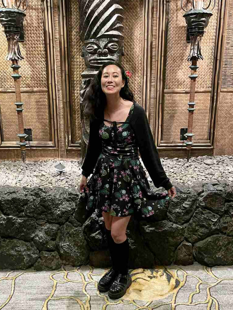 A woman posing in front of a Polynesian wall with a carved tiki figure, wearing a tiki florals kater dress with a black cardigan, black knee-high socks, and black leather shoes.