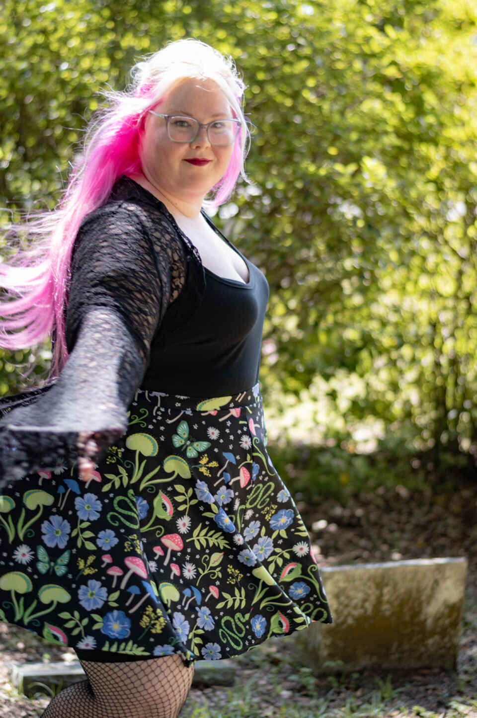 A model wearing a Halloween-themed skirt in an outdoor setting. The model wears a black top with lace sleeves and patterned skirt featuring Venus flytraps, mushrooms, and spooky florals in a lush dark botanical print.